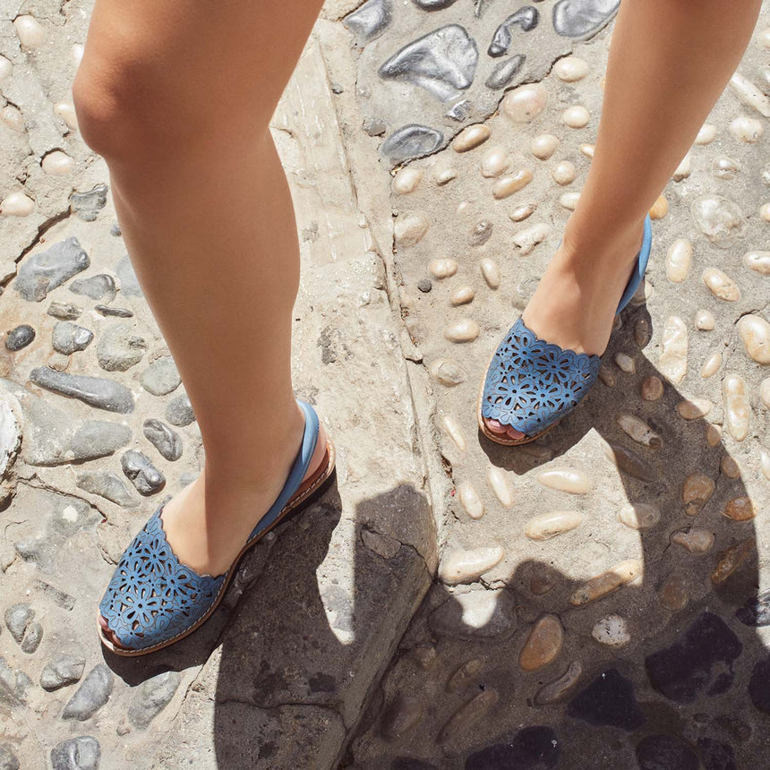 Sandalia menorquina lola Almagro con cuña y grabados de flores en la pala de la sandalia color azul, esta caminando con las sandalias por un suelo empedrado