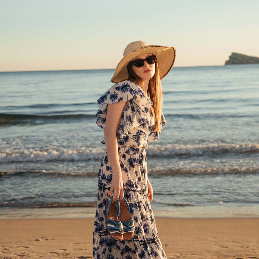 Sandalia menorquina lola Almagro planas y trenzadas con una combinación de colores blanco y azul, estas sandalias las lleva una modelo en la mano paseando por la playa